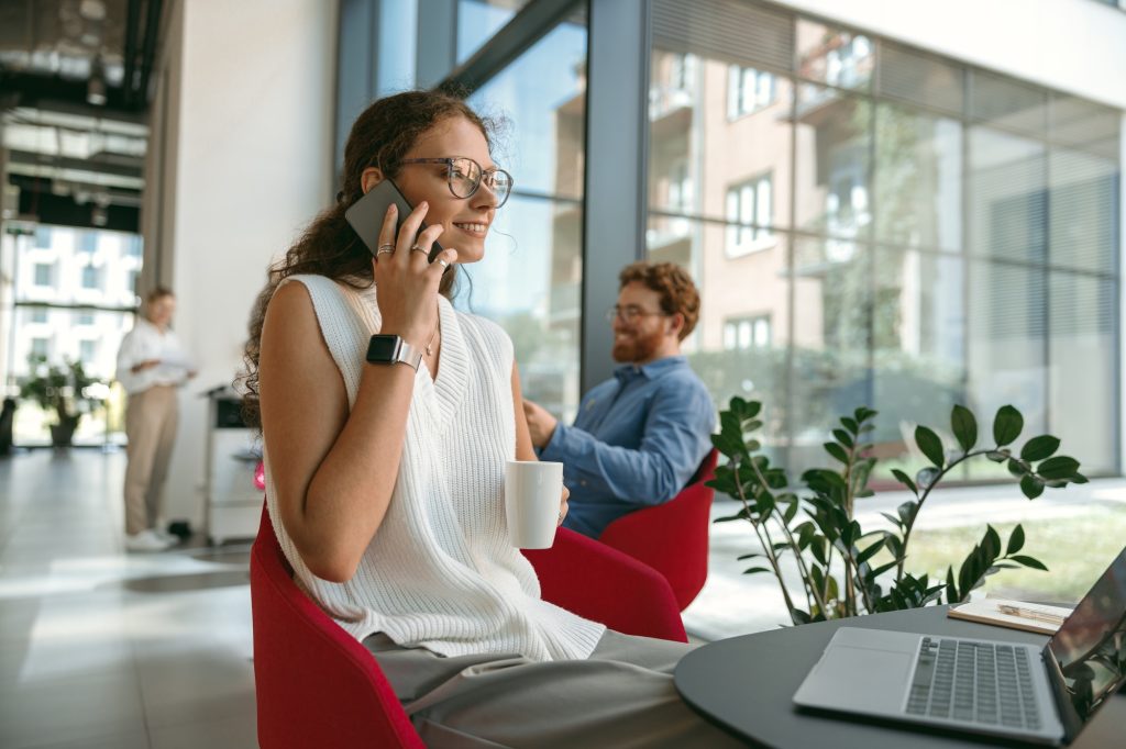 Woman freelancer talking on phone with client while working on laptop in modern coworking