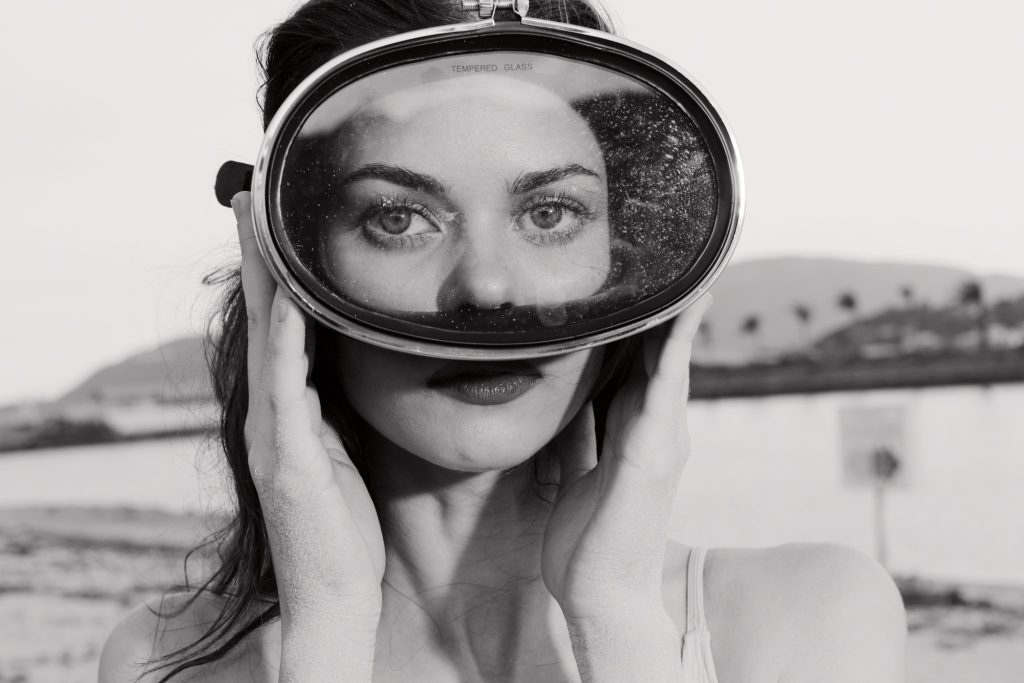 A woman holding a vintage diving mask against a blurred outdoor background Her striking blue eyes