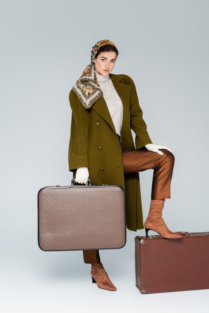 full length of trendy woman in green coat and headscarf posing with retro suitcases on grey